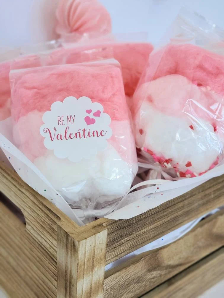 Pink and white plush toys in clear packaging with a 'Be My Valentine' label on a wooden surface.