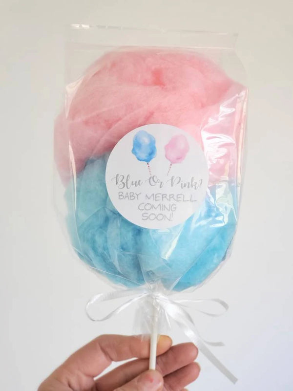 A hand holding a clear plastic bag with pink and blue cotton candy on a stick, sealed with a white ribbon and a label that reads 'Blue or Pink? Baby Gender Reveal Party!'.
